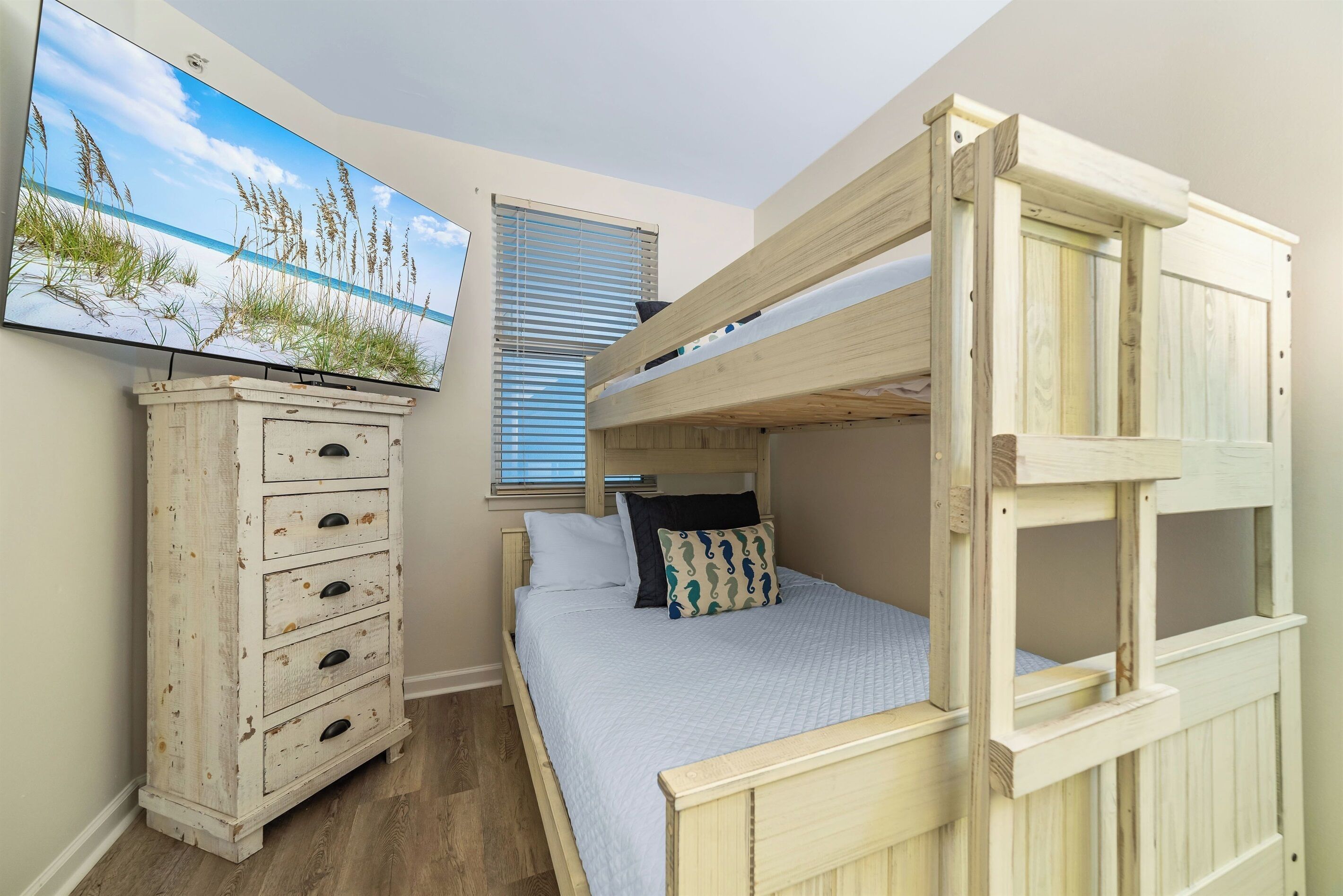 Un lit superposé avec une tête de lit, une commode avec des tiroirs et un tableau représentant de l’herbe et le ciel, accroché au mur.