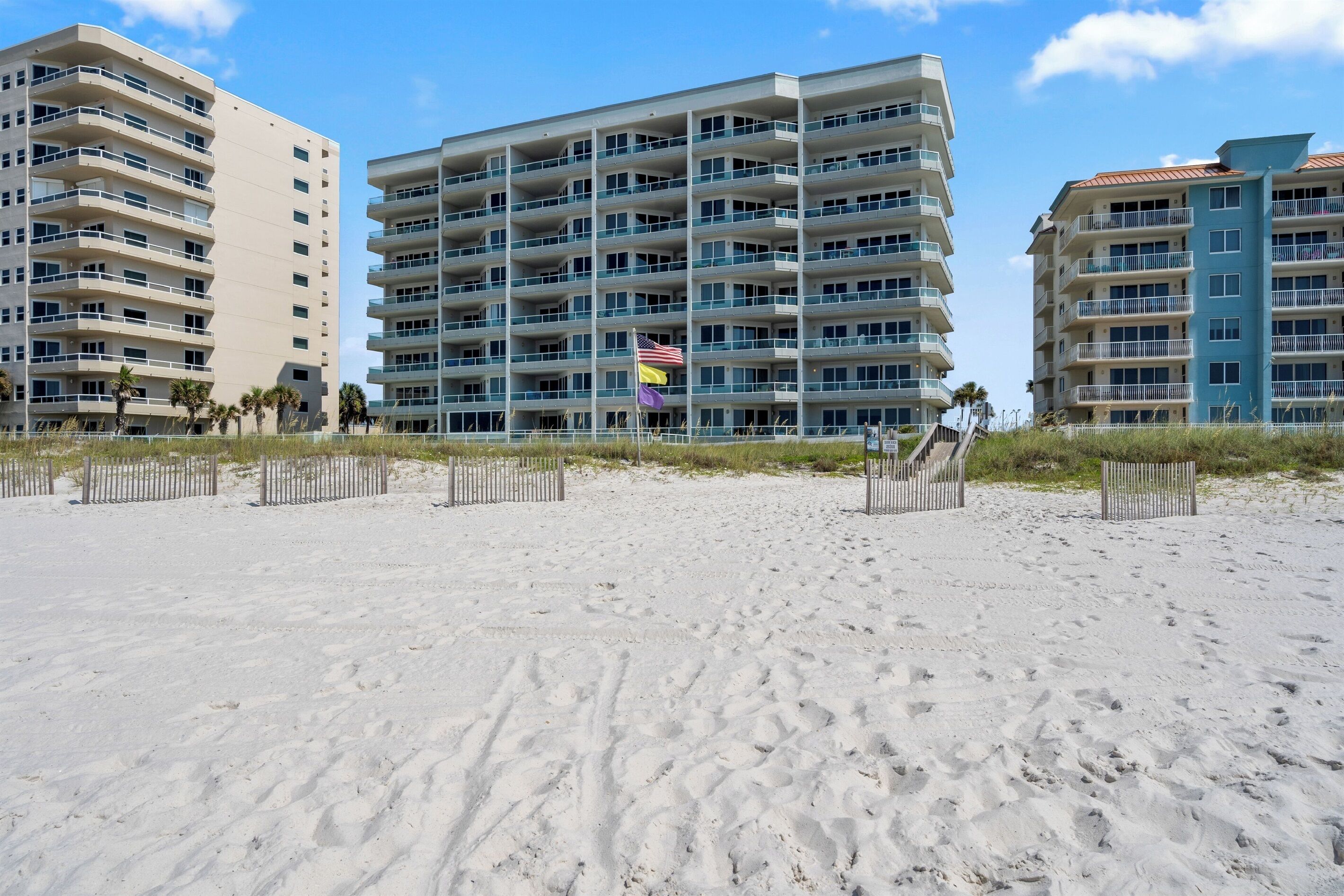 Ein Strandhotel mit mehreren Balkonen, ein Sandstrand im Vordergrund und ein klarer Himmel.