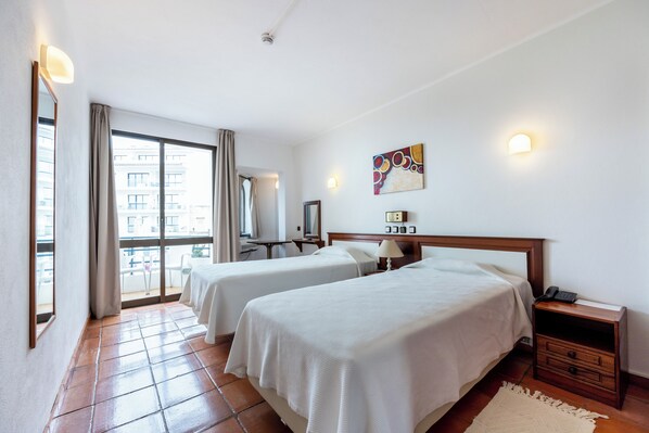Habitación de hotel con dos camas, una mesita de noche, una lámpara, un cuadro y vistas al balcón exterior.