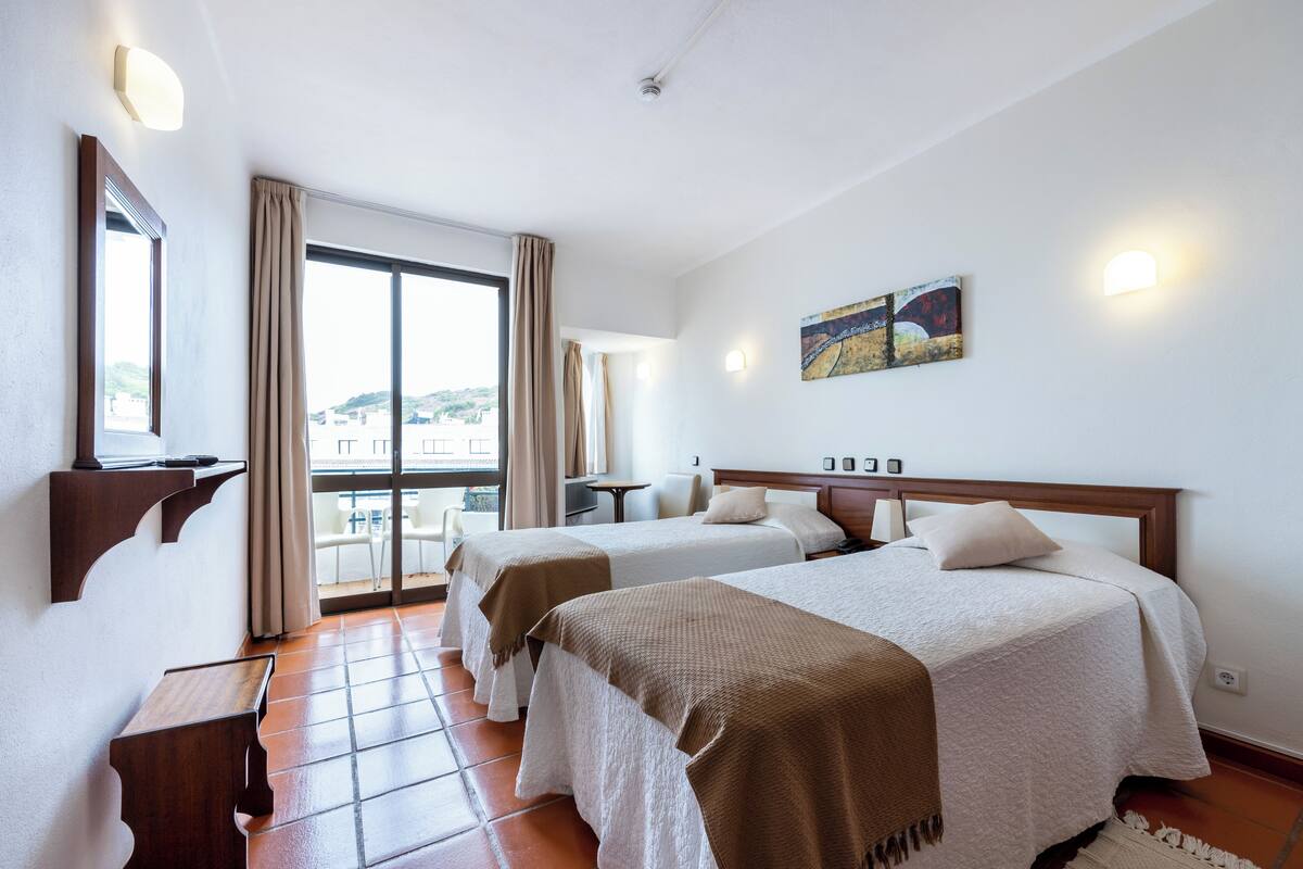 A hotel room with two beds, a large window with a view, a painting on the wall, and a small table.