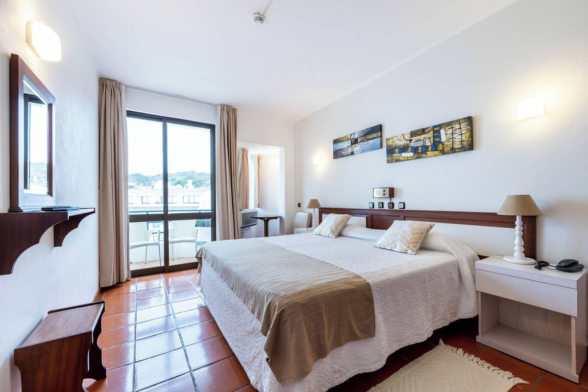A hotel room with a large bed, a desk, a chair, a nightstand, and a view of the outdoors.