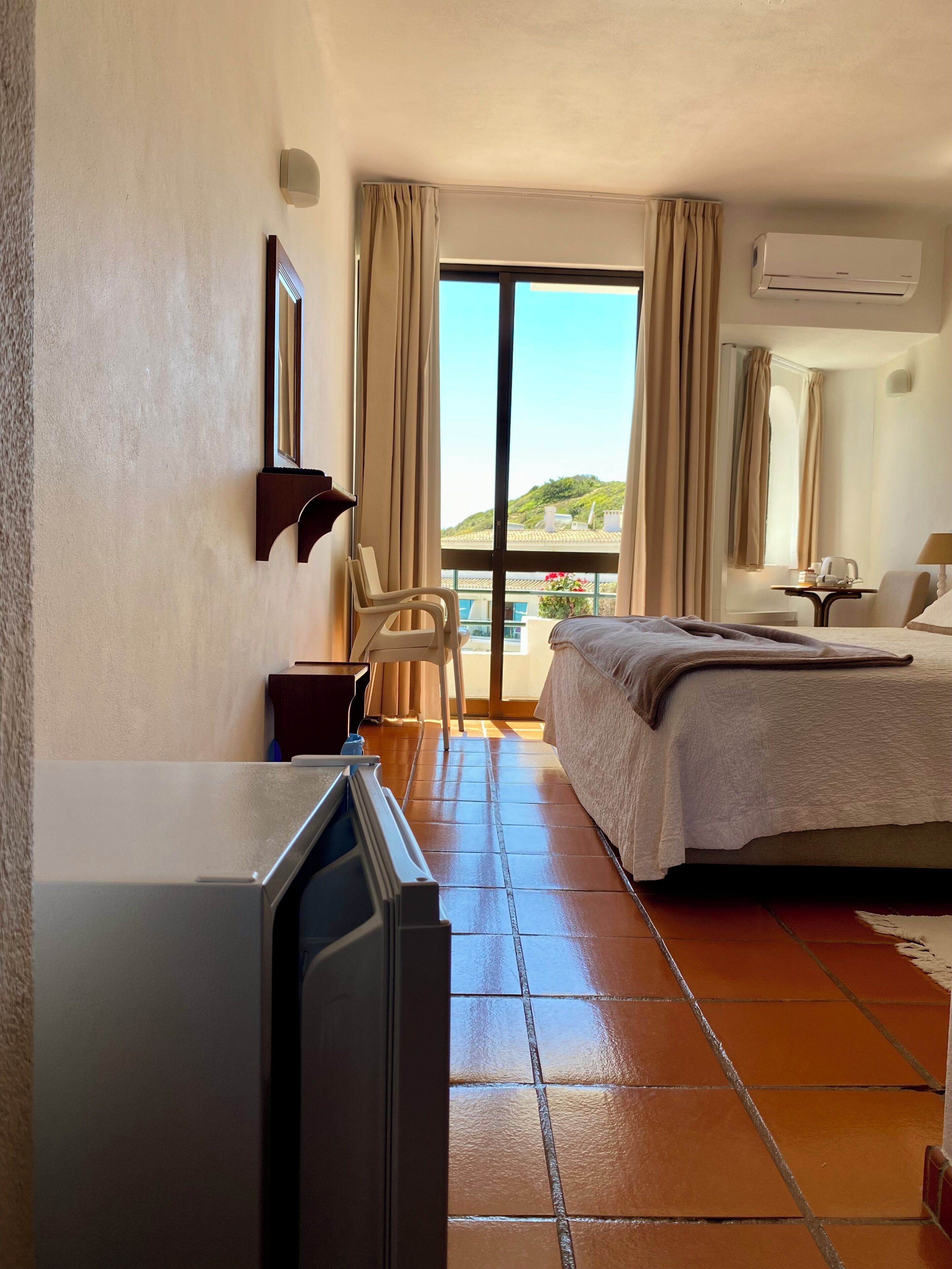 Habitación de hotel con cama, escritorio, silla, televisor y balcón con vistas a las colinas.