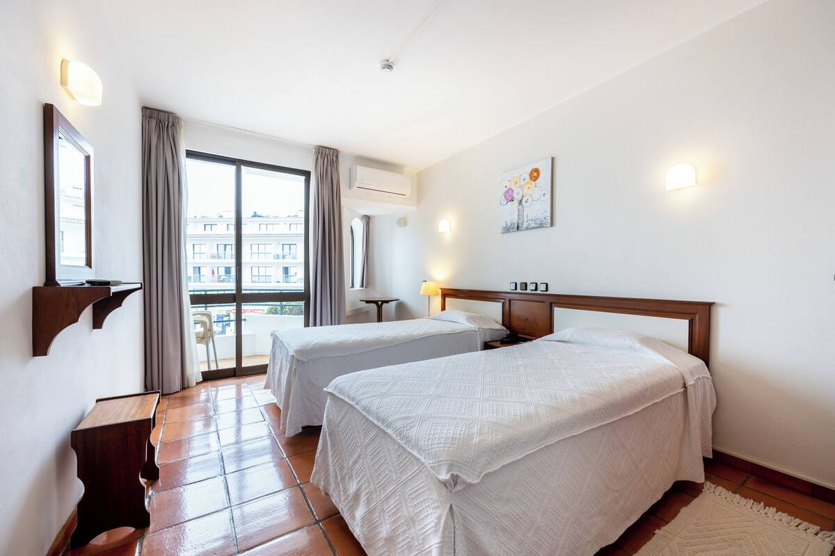 A hotel room with two beds, a balcony with a view, a small table, and a wall-mounted air conditioner.