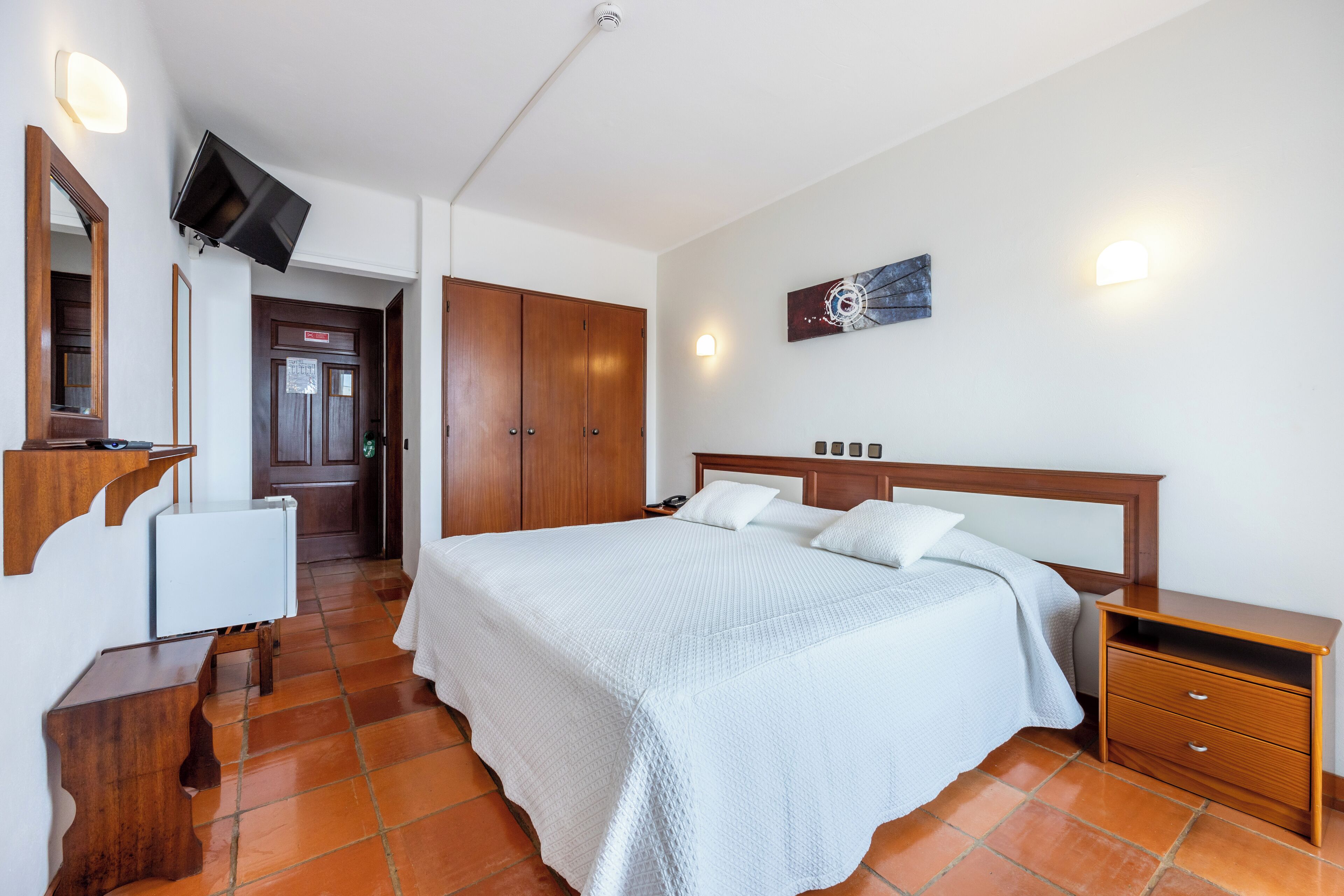 A bedroom with a large bed, wooden wardrobe, a bedside table, a TV mounted on the wall, and a door leading to another room.