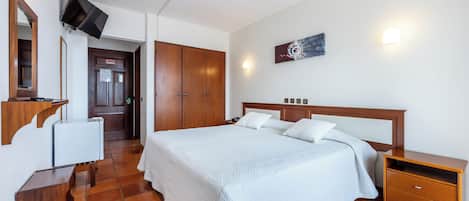 A bedroom with a large bed, wooden wardrobe, a bedside table, a TV mounted on the wall, and a door leading to another room.