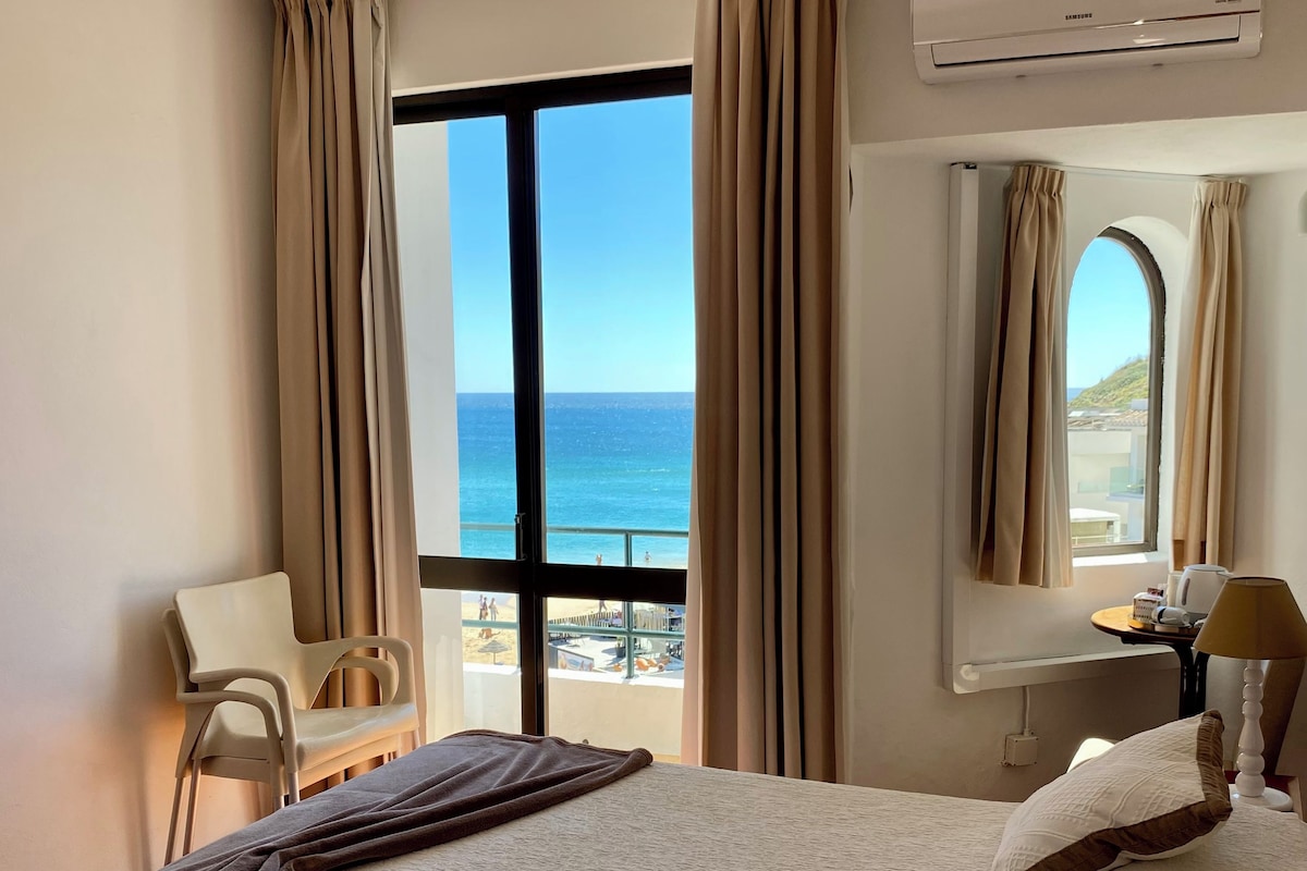 Habitación de hotel con cama, una silla, lámpara, equipo de aire acondicionado y vista al mar.