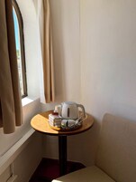 Um canto do quarto com uma janela, uma mesinha com uma chaleira, uma xícara e uma caixa de lenços.