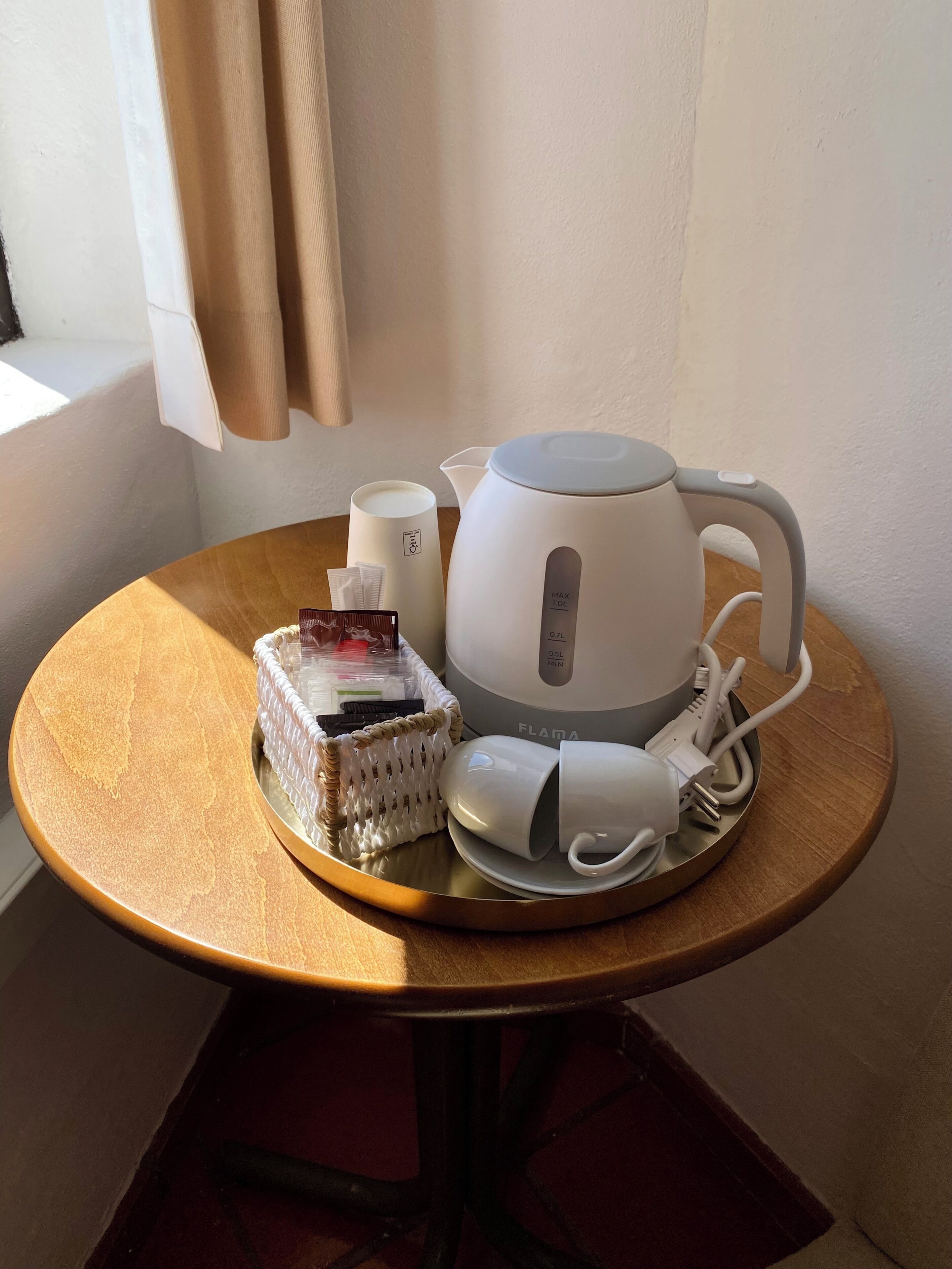 Una mesa con un hervidor blanco, una taza blanca, un cargador blanco y una cesta tejida con objetos dentro.