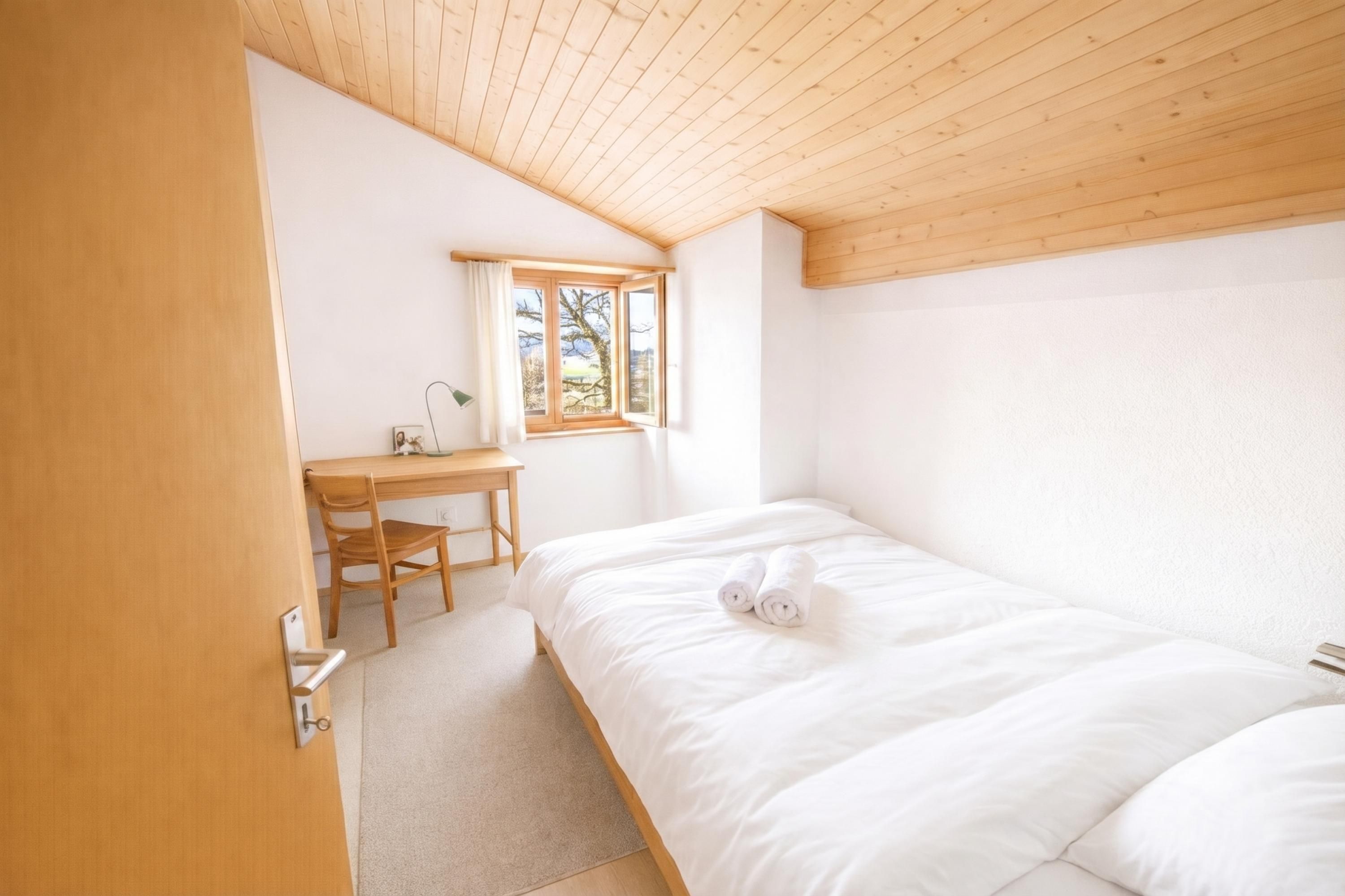 A bedroom with a wooden ceiling, a bed with white bedding, a desk with a chair, and a window.