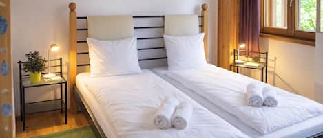 A double bed with white linens and rolled towels, two bedside tables with lamps, a green rug, and a window with curtains.