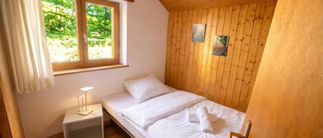 A single bed with white linens, a bedside table with a lamp, a window with curtains, and wooden paneling on the walls.