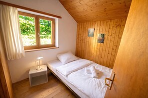 Un lit simple avec du linge de lit blanc, une table de chevet avec une lampe, une fenêtre avec des rideaux et des lambris en bois sur les murs.