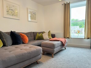 Ein Wohnzimmer mit einem grauen Ecksofa, einem grauen Hocker und einem Fenster mit Vorhängen.
