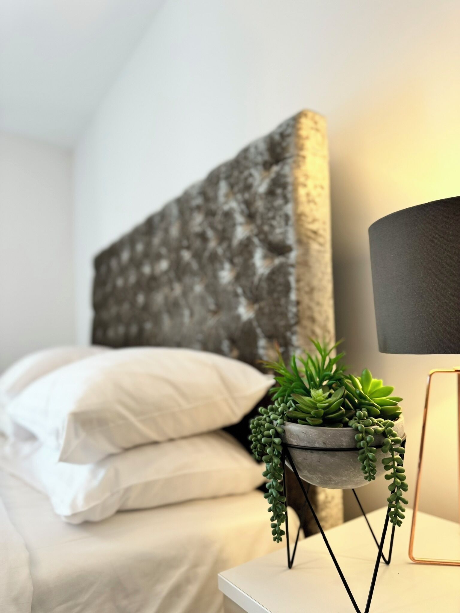 A neatly made bed with white linens and a grey headboard. A small potted plant with green foliage is placed on a white bedside table next to a lamp with a black shade.