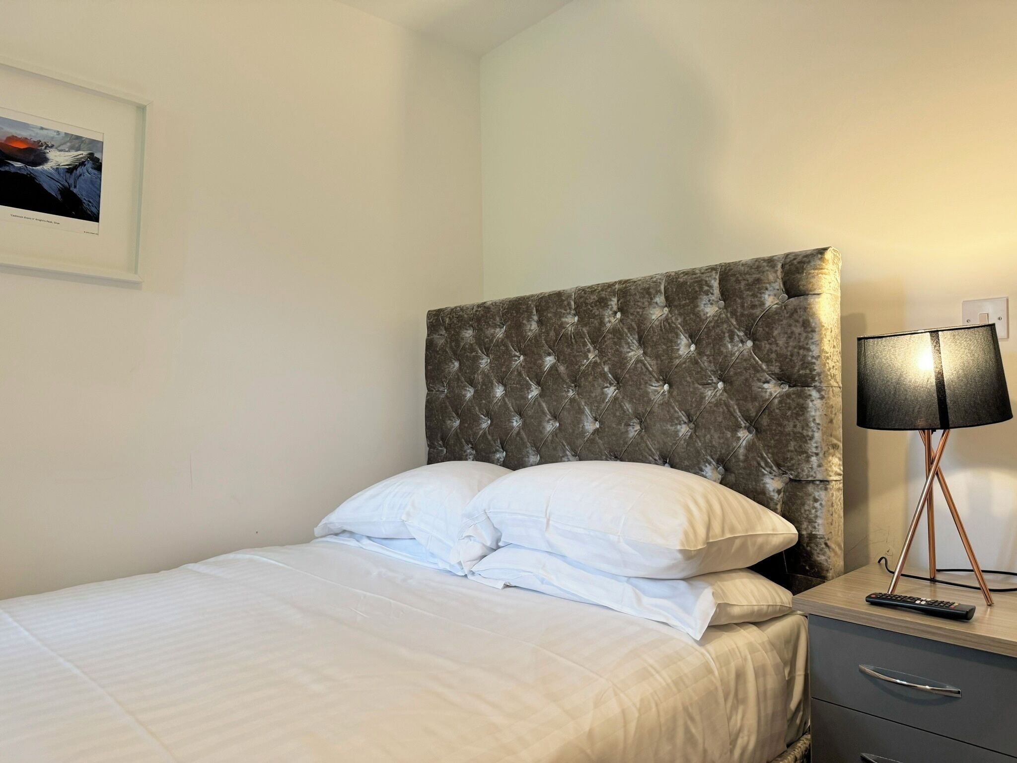 A neatly made bed with white linens, a tufted headboard, a bedside table with a lamp, and a remote control.