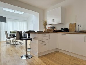 A modern kitchen with white cabinets, a wooden countertop, and a bar stool. There is a dining area with a table and chairs visible in the background. - The Courtyard, 4 - Bed, Sleeps 6, Pets Welcome (Ulverston)