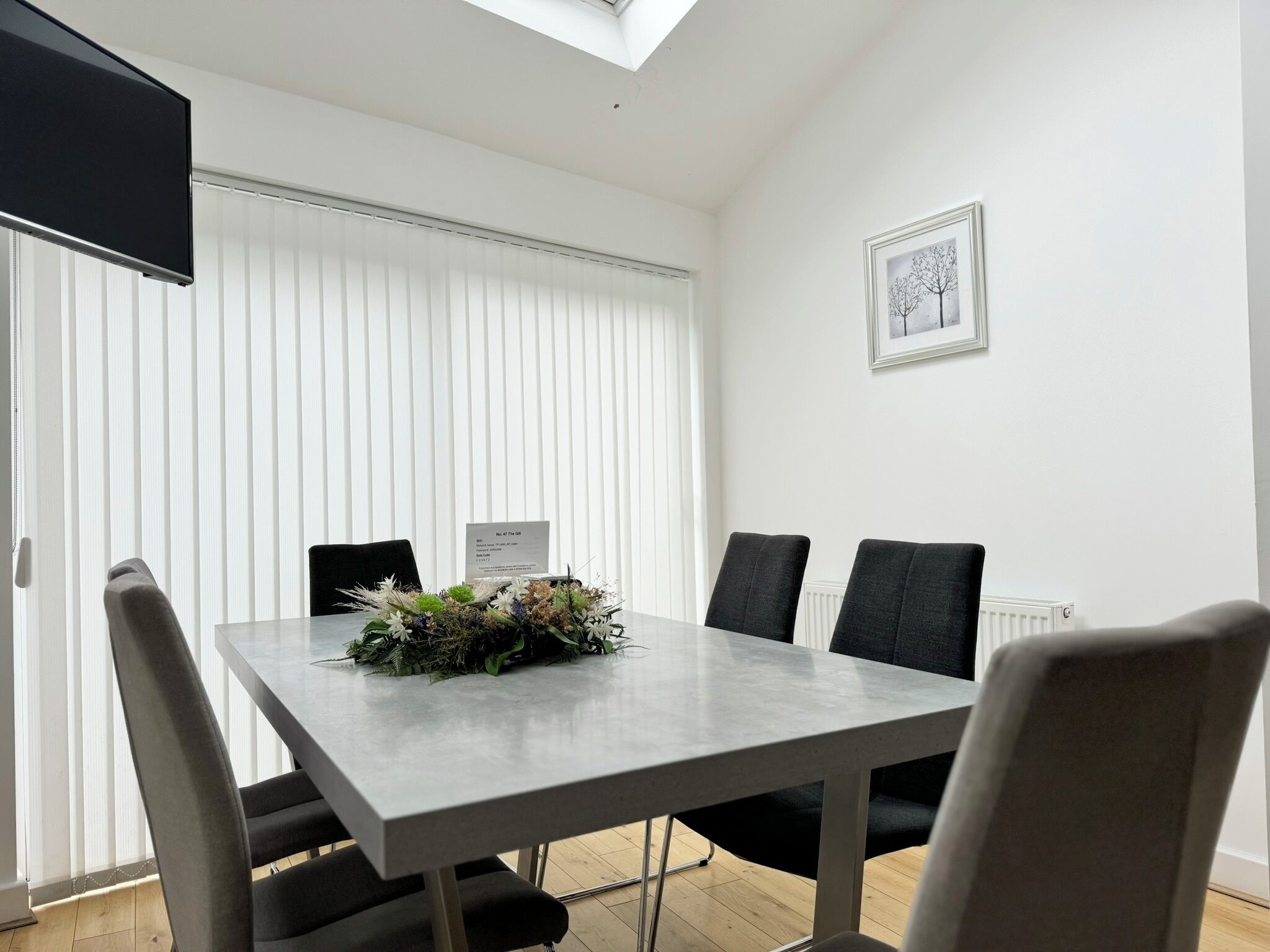 A dining area with a table, chairs, a flower arrangement, a framed picture, and a TV on the wall.