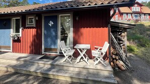 A red wooden cabin with a blue door, two white folding chairs, and a small table on a wooden deck. - Room-archipelago Cottage is Utö (Uto)
