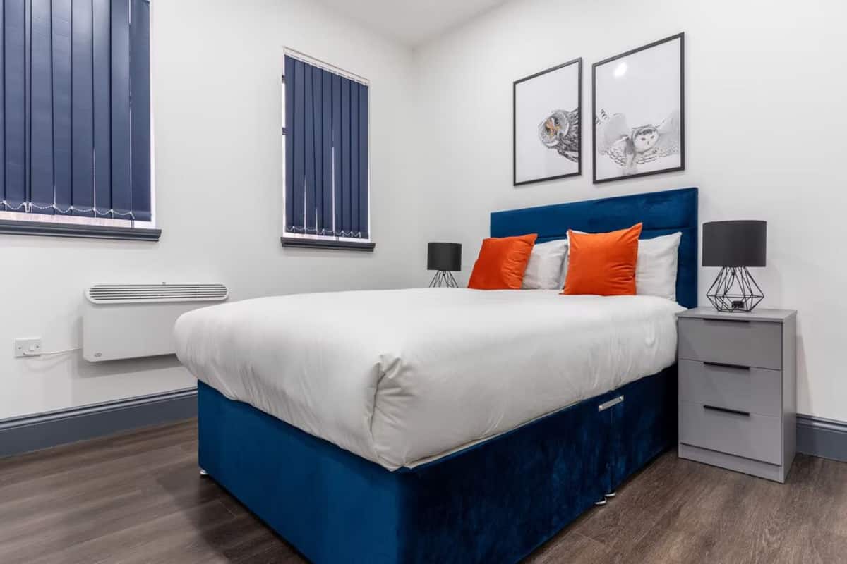 Une chambre moderne avec un lit bleu, une literie blanche et des coussins orange. Deux tableaux encadrés ornent le mur, il y a une fenêtre avec des stores et une table de chevet avec une lampe.