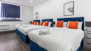 Een hotelkamer met twee bedden, elk met blauwe hoofdborden en witte beddengoed. Er zijn oranje kussens, een ingelijste foto aan de muur en een raam met gordijnen.