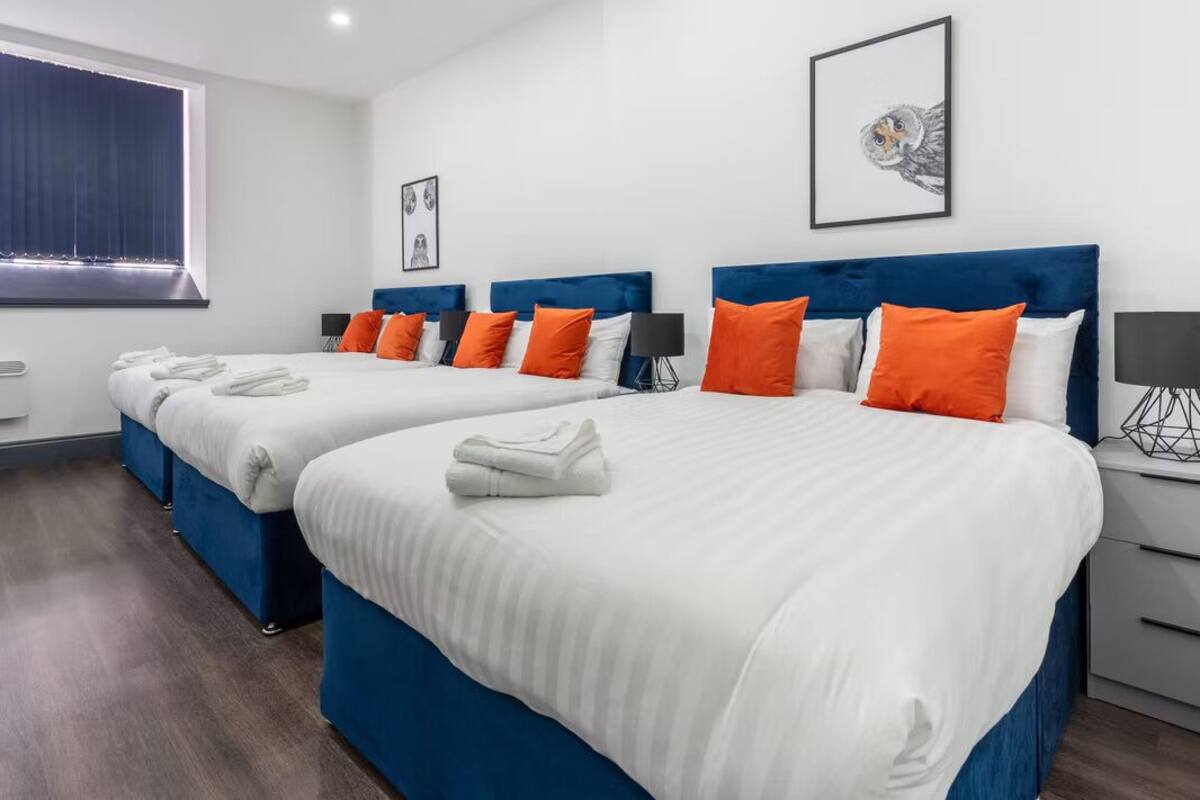 Une chambre d’hôtel avec deux lits, chacun doté d’une tête de lit bleue et de draps blancs. On y trouve des oreillers orange, un tableau encadré au mur et une fenêtre avec des rideaux.