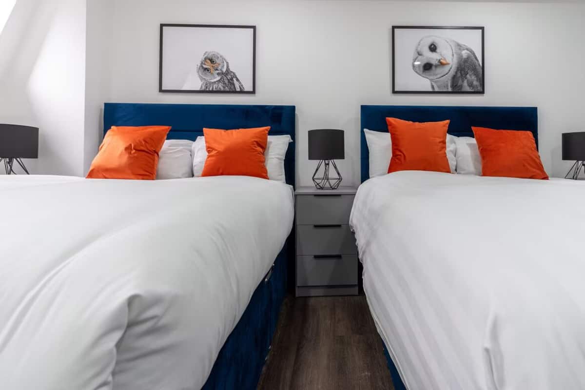 Une chambre d’hôtel avec deux lits, chacun doté de draps blancs et d’oreillers orange, d’une tête de lit bleue et d’une table de chevet avec une lampe.