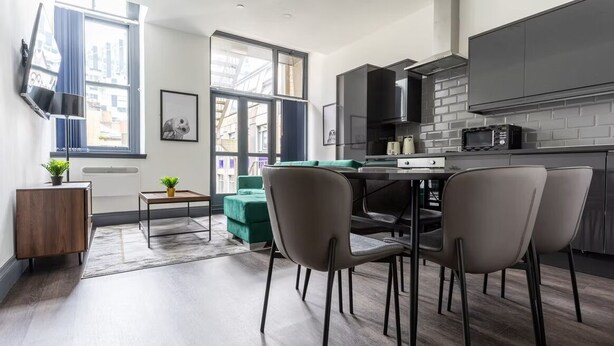 Een moderne keuken met donkere kastjes, een eethoek met een tafel en stoelen, en een stadszicht door grote ramen.