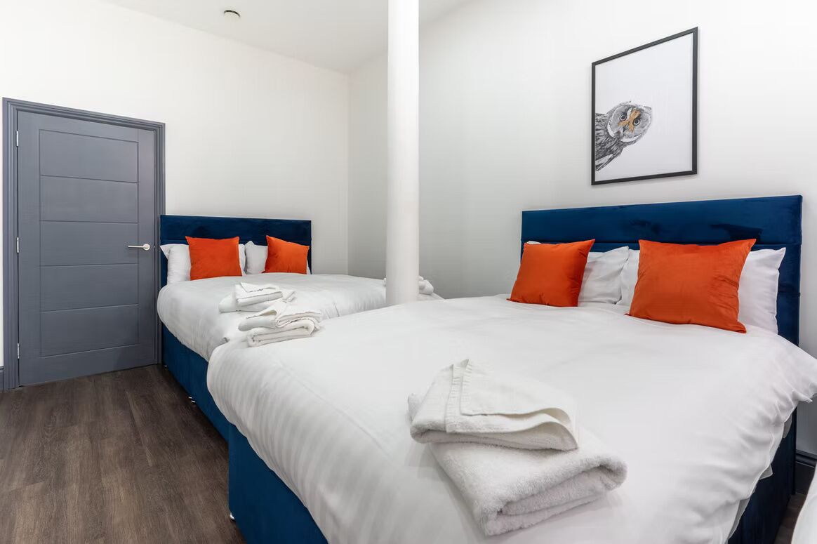 Ett hotellrum med två sängar, vita sängkläder, orange kuddar, ett inramat konstverk med ett ugglemotiv på väggen och en grå dörr.
