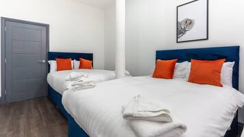 Ett hotellrum med två sängar, vita sängkläder, orange kuddar, ett inramat konstverk med ett ugglemotiv på väggen och en grå dörr.