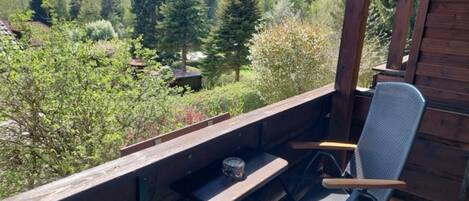 A wooden deck with a metal chair and a table, overlooking a forested area with mountains in the distance.