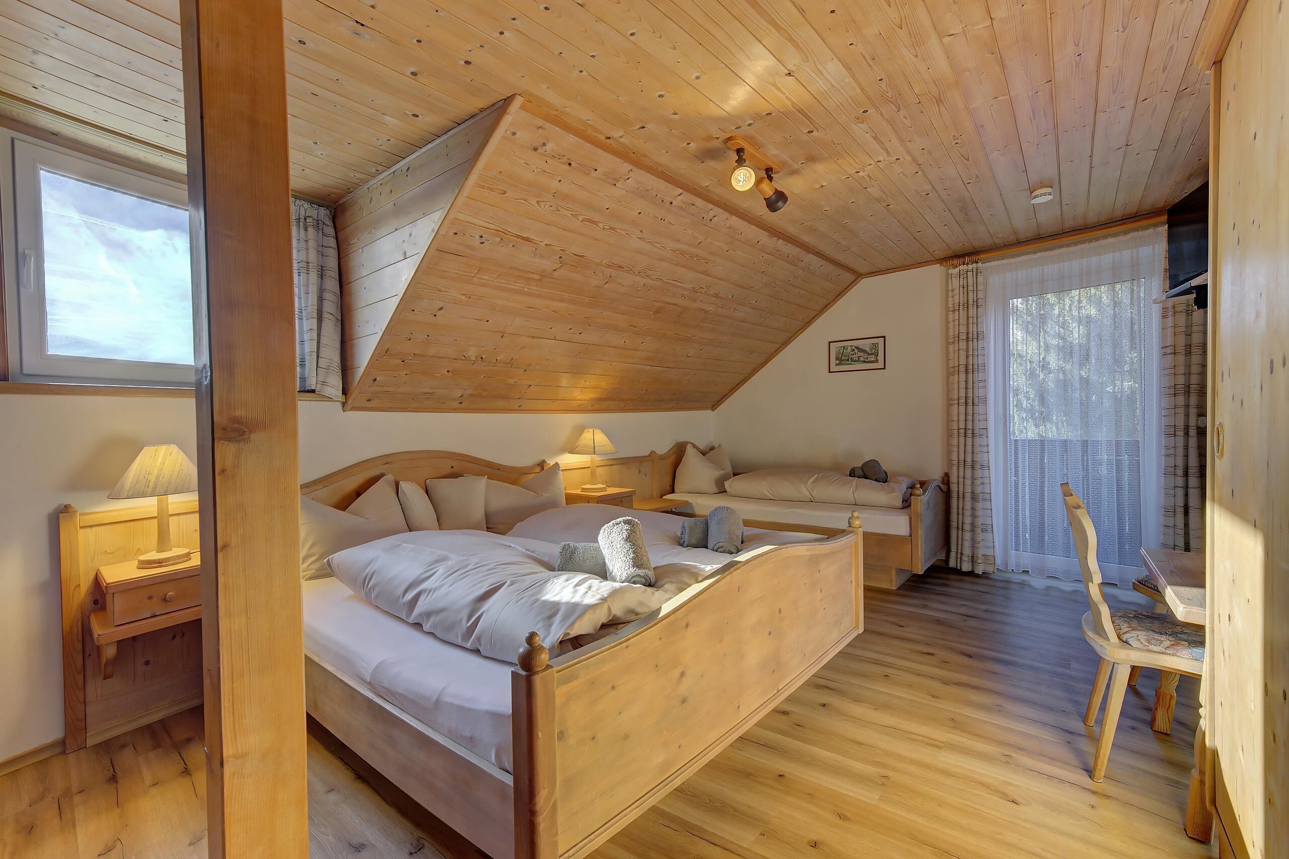 A bedroom with a wooden ceiling, a bed, a bedside table with a lamp, a chair, and a window with curtains.