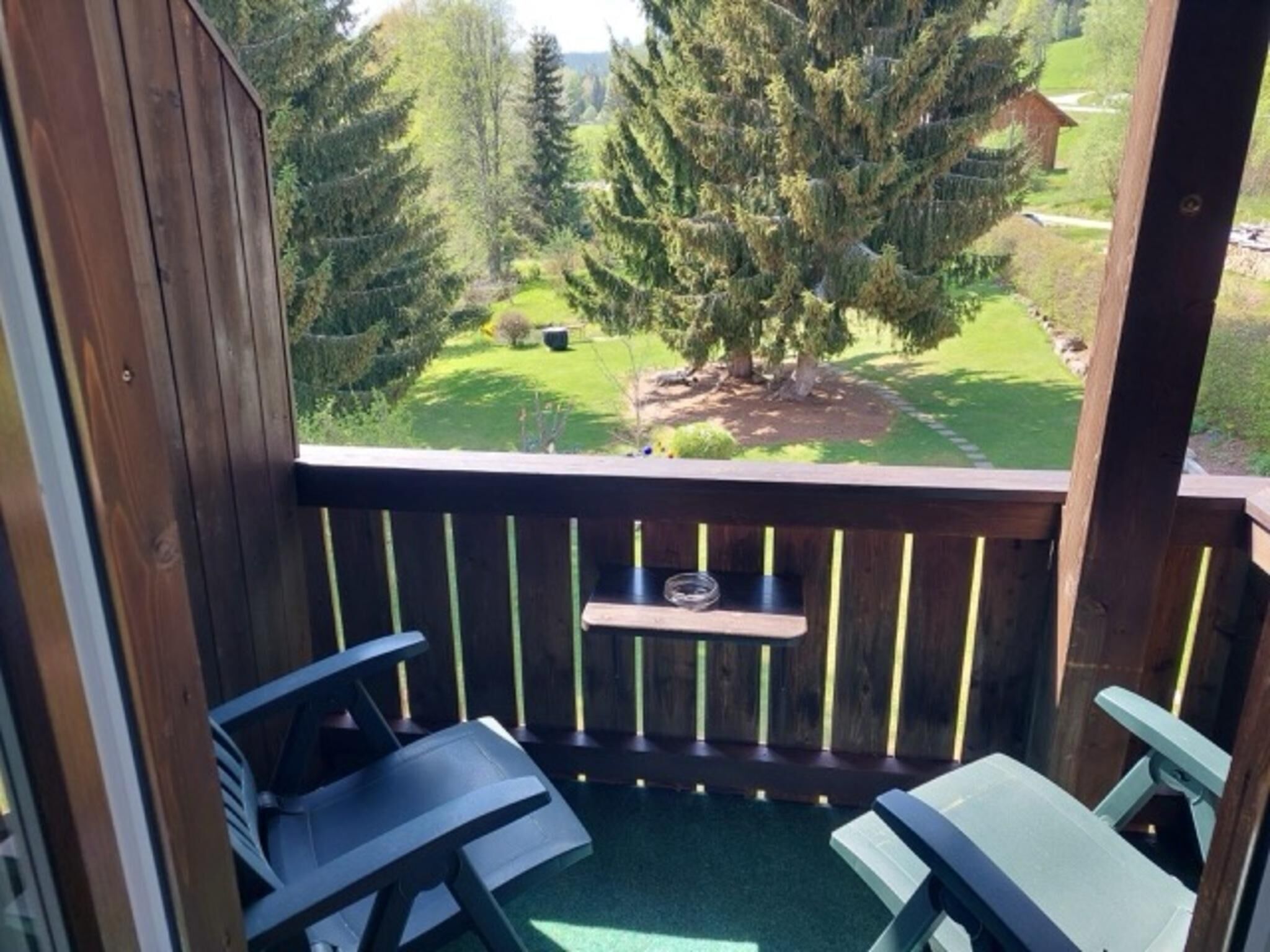 Ein Holzbalkon mit zwei Stühlen und einem kleinen Tisch, mit Blick auf einen gepflegten Garten mit einem großen Baum und einer Rasenfläche.