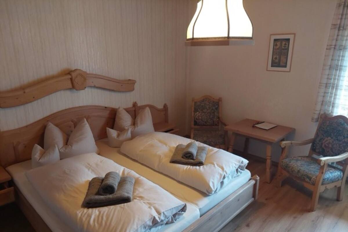 Ein Doppelbett mit Holz-Kopfteil, ein Schreibtisch, ein Stuhl und ein Fenster mit Vorhängen.