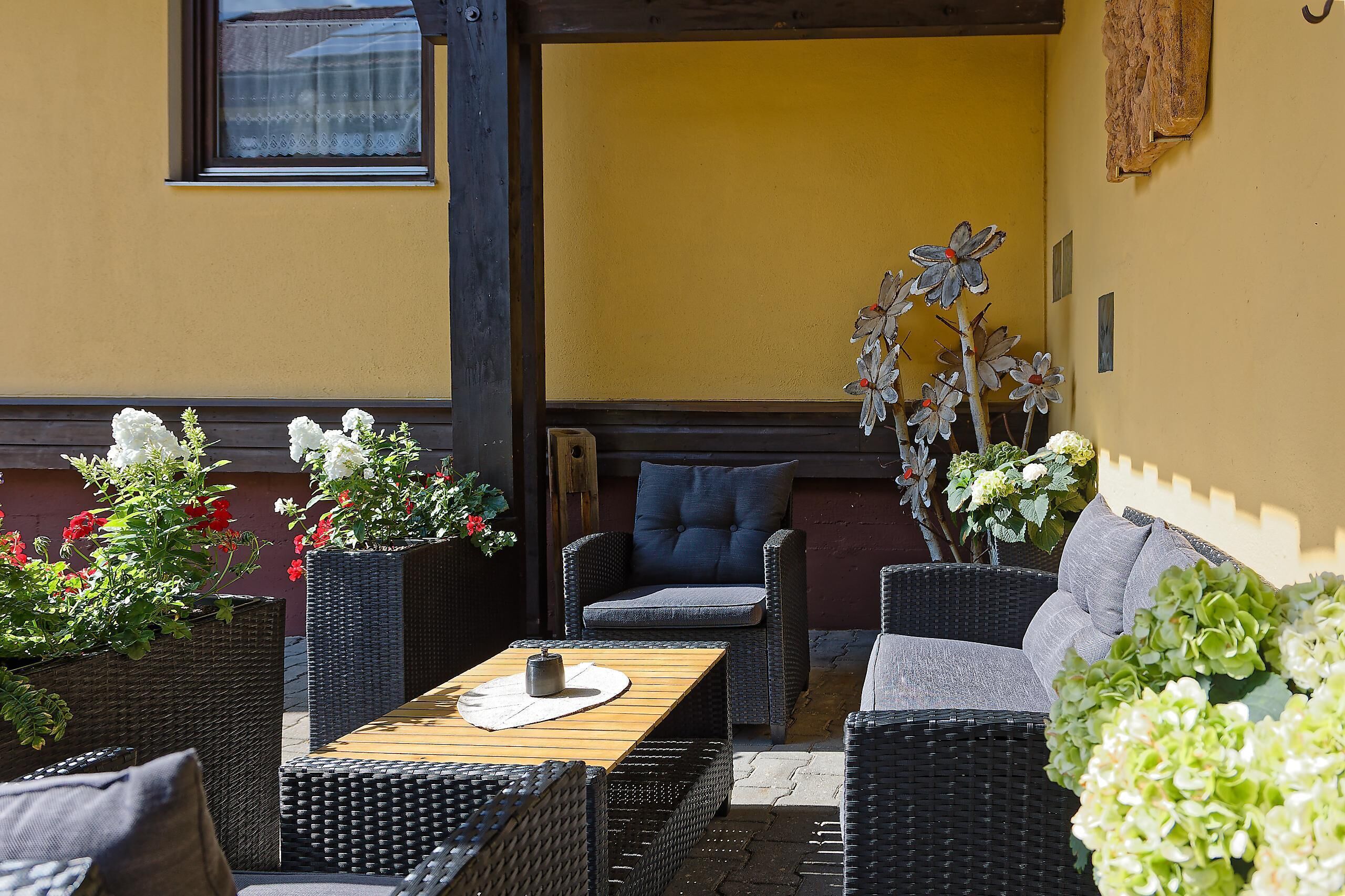 Ein Sitzbereich im Freien mit Korbmöbeln und einem Tisch, umgeben von Blumentöpfen und Blumen.