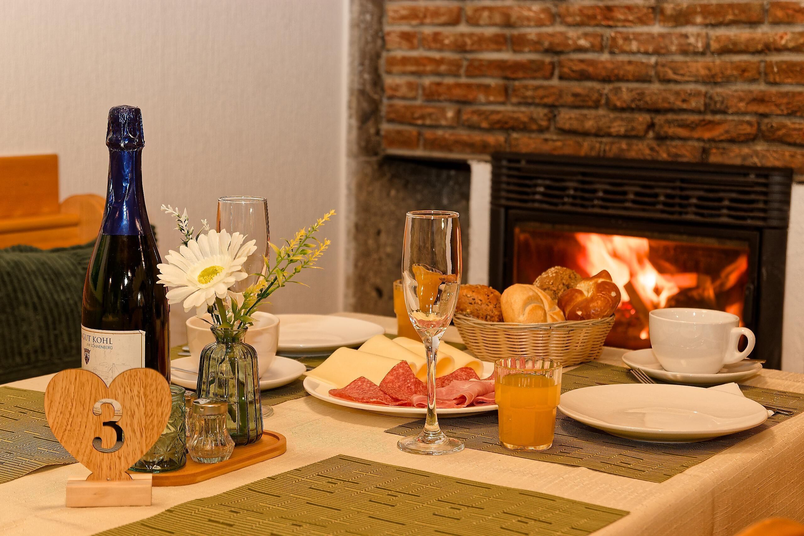Morgenmadsbord med flaske, glas, blomster, brød og tallerkener nær en pejs.