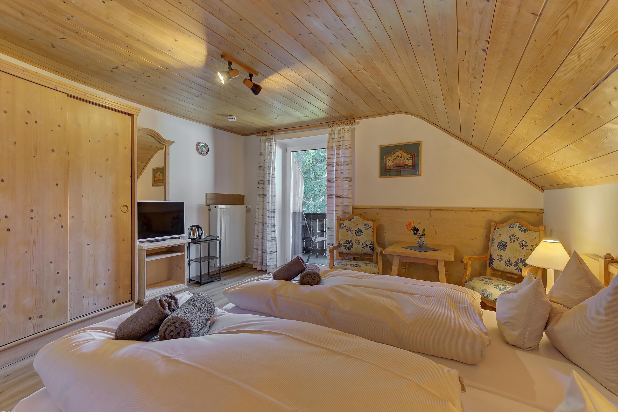 A cozy bedroom with a wooden ceiling, a bed with white linens, a desk with a computer, and a chair with a floral cushion.