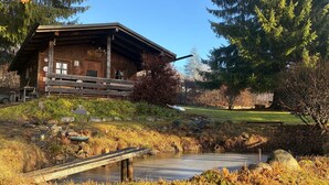 Une cabane en bois rustique, dotée d’un porche, entourée d’un étang et d’une zone herbeuse parsemée d’arbres.