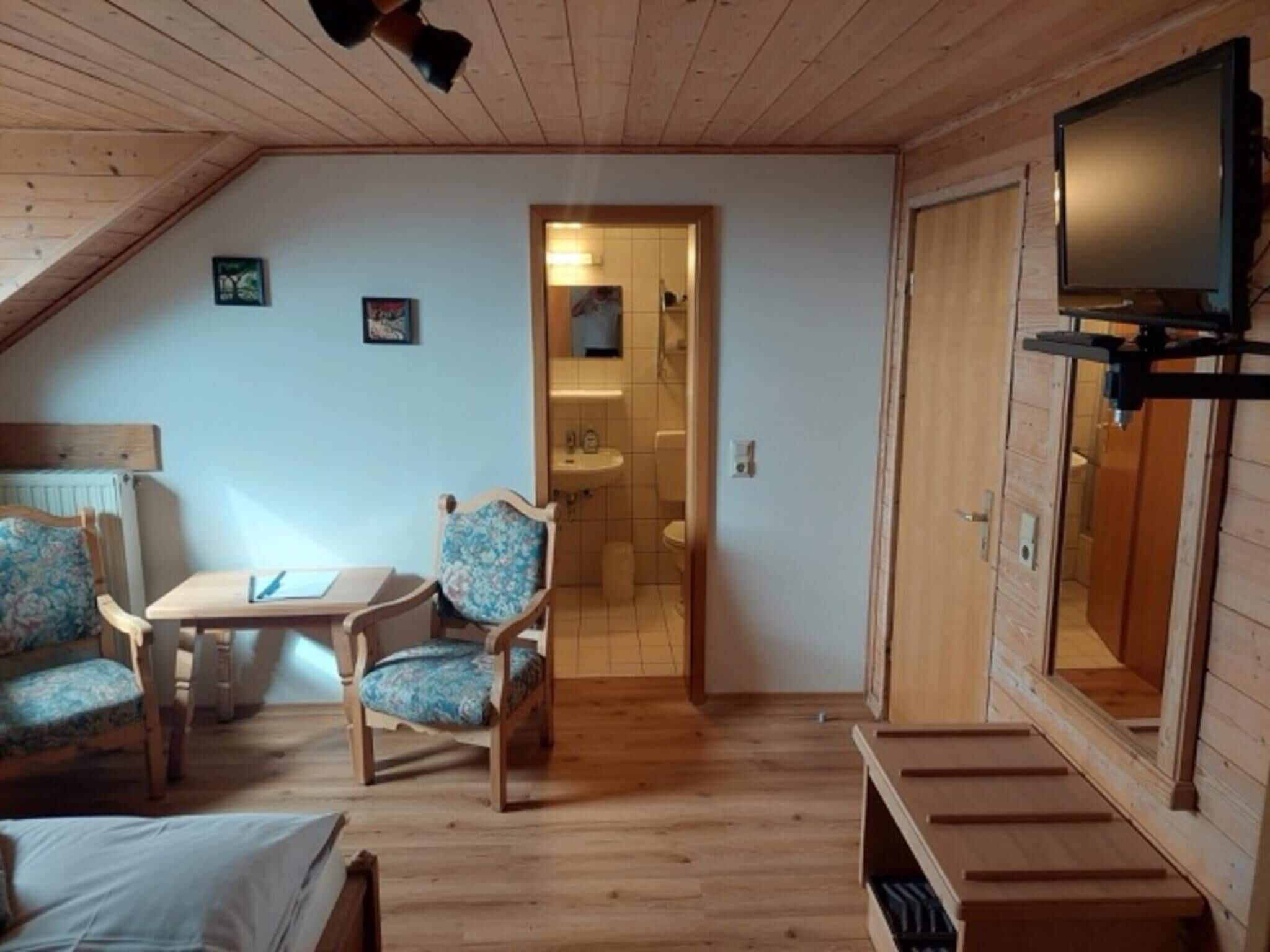 Una habitación acogedora con techo de madera, un televisor en la pared, una pequeña mesa con sillas y una puerta que da al baño.