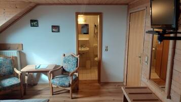 Una habitación acogedora con techo de madera, un televisor en la pared, una pequeña mesa con sillas y una puerta que da al baño.