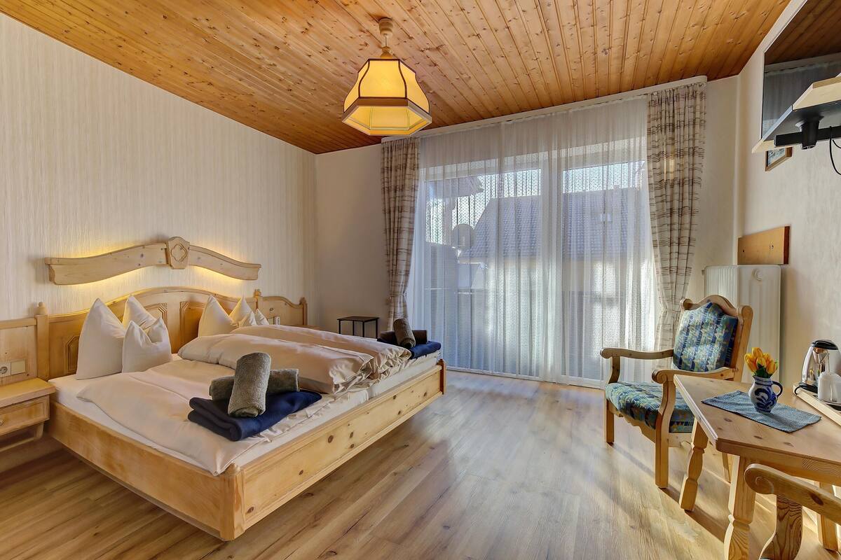 Ein Bett mit Holzrahmen, Kissen und Decke, ein Holztisch mit einer Vase und ein Stuhl mit Sitzkissen.