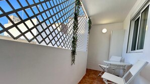Ein Balkon mit weißen Wänden, einem weißen Tisch und Stuhl und Blick auf Gebäude durch ein Gitter.