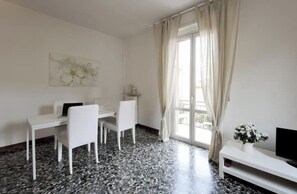 Ein Zimmer mit weißem Schreibtisch, zwei Stühlen, einem Fernseher auf einem Standfuß und einem Fenster mit Vorhängen.