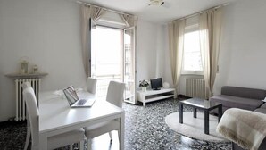 Ein modernes Wohnzimmer mit einem weißen Sofa, einem Esstisch mit einem Laptop, einem Fernseher auf einem Standfuß und einem Deckenventilator.