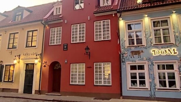 A cobblestone street with buildings featuring traditional European architecture, including a red building with a shop sign and a blue building with a 'DOM' sign. - Riga Stylish Old Town getaway next to Dome square (Rīga)