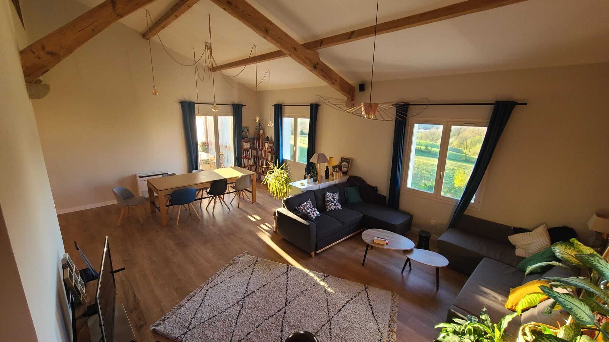 Een ruime woonkamer met een hoge plafond, houten balken, een eettafel met stoelen, een bank, een klein bijzettafeltje en een groot raam met uitzicht op de omgeving.