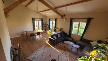 Een ruime woonkamer met een hoge plafond, houten balken, een eettafel met stoelen, een bank, een klein bijzettafeltje en een groot raam met uitzicht op de omgeving.