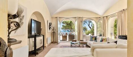 A spacious living room with a large bed, a television, and a view of the outdoors through arched windows.