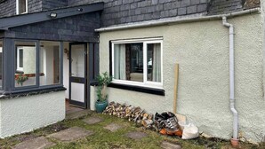 A two-story house with a dark roof, white exterior walls, and a small garden area with a wooden fence and a potted plant. - Hot tub - Garden - Bbq - Parking - Pets - Sleeps 6 (Strathpeffer)