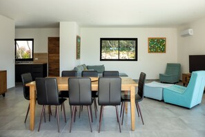 Ein modernes Wohnzimmer mit einem Holzesstisch, schwarzen Stühlen, einem petrolfarbenen Sofa und einem großen Fenster mit Blick auf Bäume.