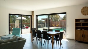 Amplia sala de estar con mesa de comedor, sillas y sofá. Una gran puerta corrediza de vidrio da a un patio exterior con piscina y mobiliario.